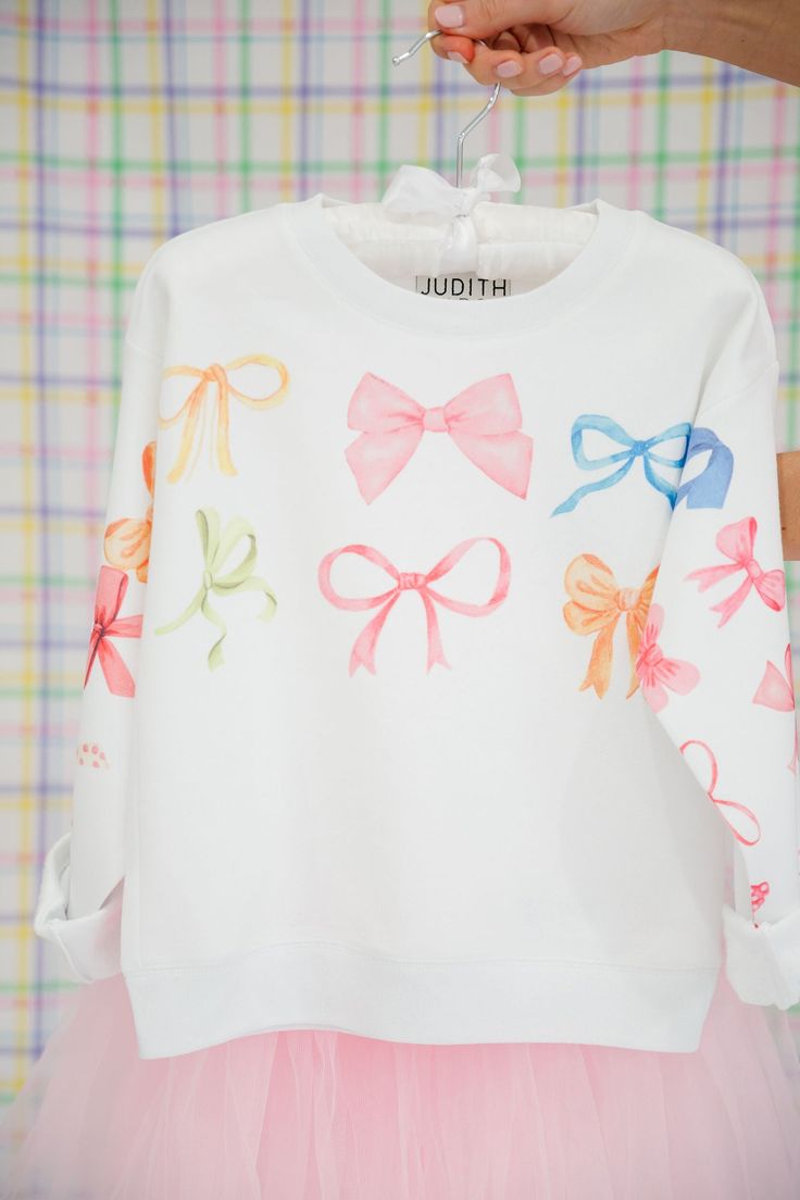 Perfect for Easter egg hunts and springtime adventures, our MINI KIDS BOW PRINT PULLOVER adds a pop of playful style to any outfit. The white pullover features a charming print of pastel colored bows, making it a delightful addition to your little one's wardrobe. Time to hop to it! This is a made-to-order item. All customized orders are currently shipping within 14 business days. To receive item quicker, expedited shipping is available at checkout. Cute Pastel Tops For Spring, Casual Tops With Bow Print For Spring, Casual Bow Print Tops For Spring, Spring Crew Neck Top With Bow Print, Cute White Top With Bow, Cute White Top With Bow Detail, Spring Bow Print Crew Neck Top, Playful Pastel Tops For Spring, White Cotton Top With Pink Bow