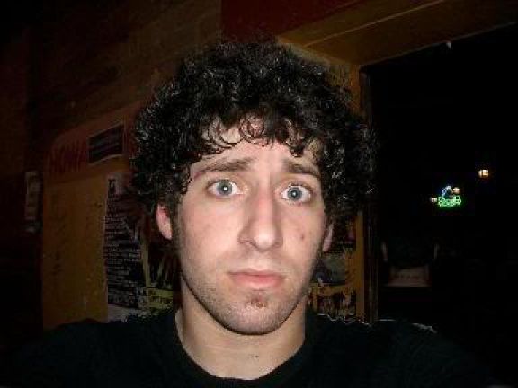 a young man with curly hair and blue eyes looks at the camera while wearing a black t - shirt