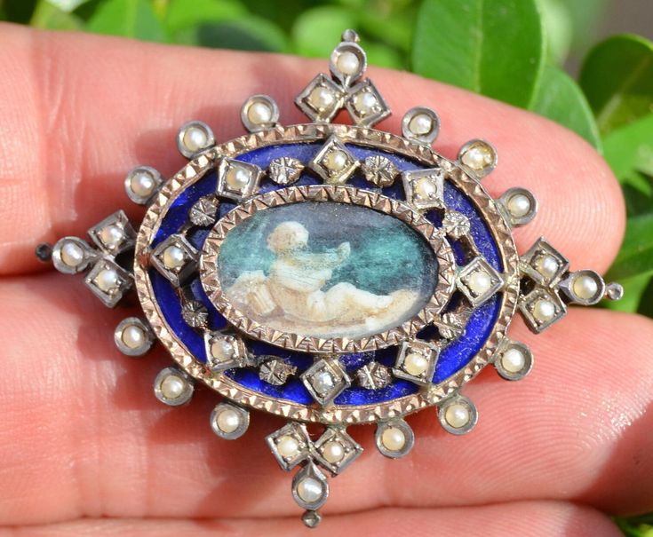 Adorable hand painted enamel brooch. This brooch is crafted from 14K as well as sterling silver. It features natural seed pearls set through out. The center of this brooch features a hand painted cherub playing an instrument. It has been set behind glass with an etched silver frame. Surrounding the portrait is fine royal blue enamel which further accents the painting. The enamel is adorned with alternating silver bits and seed pearls individually framed and set. The outer most portion is additio Wedding Brooches, Micro Mosaic Jewelry, Mosaic Jewelry, Straight Pins, Wedding Brooch, Micro Mosaic, Enamel Brooch, Pearl Set, Seed Pearl