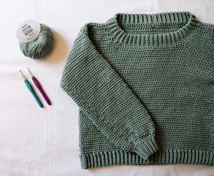 a green sweater next to two crochet hooks and a ball of yarn on a white surface