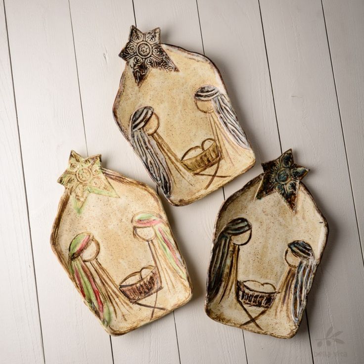 three ceramic ornaments are sitting on a white wooden surface, one has an angel and the other is a nativity scene