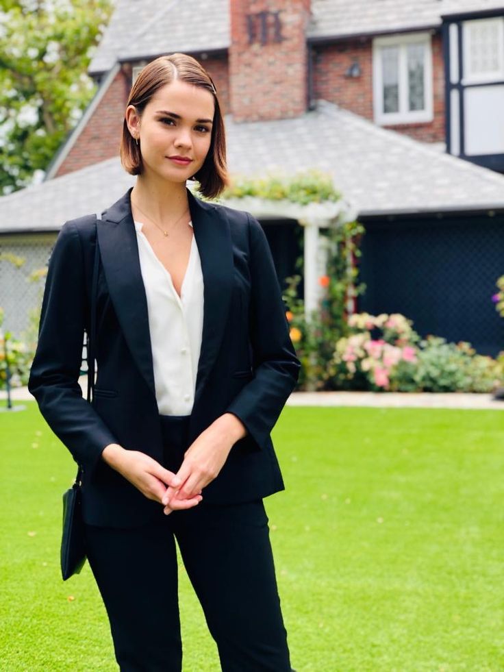 a woman standing in the grass wearing a black suit and white shirt with her hands in her pockets