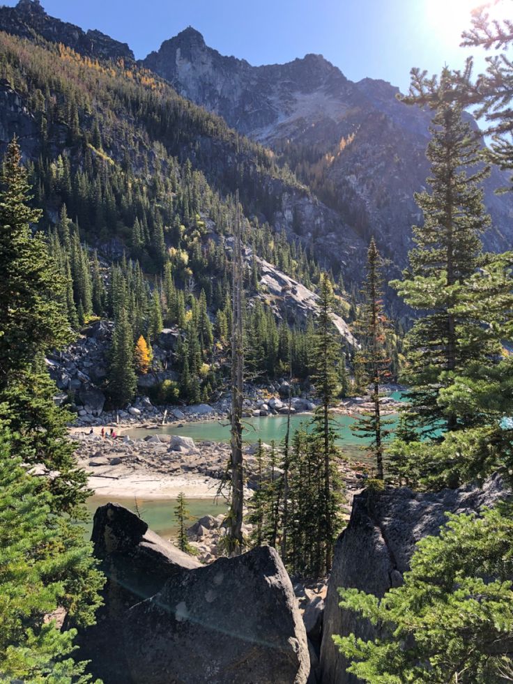 the sun shines on some trees and rocks in front of a mountain lake that is surrounded by mountains