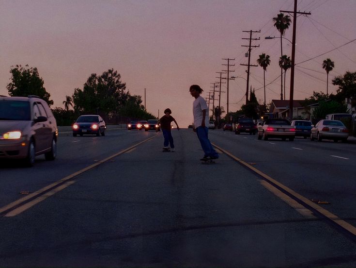 two people riding skateboards down the middle of a street at dusk with cars parked on both sides