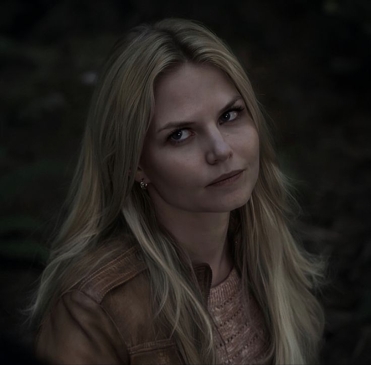 a woman with long blonde hair and blue eyes looks into the camera while wearing a brown jacket