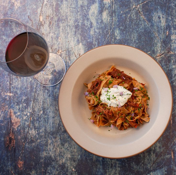 a white plate topped with pasta and sauce next to a glass of wine on top of a wooden table