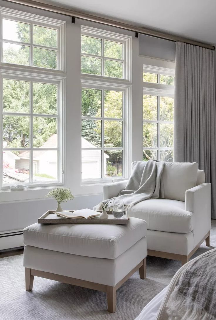 a white chair and ottoman in front of two large windows with drapes on them