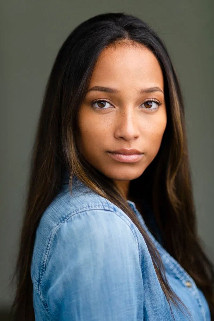 a woman with long brown hair wearing a denim shirt and looking at the camera while standing in front of a gray wall