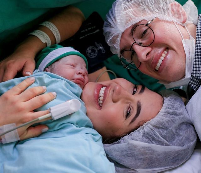 two women smile as they hold a baby in a hospital gown and headgear