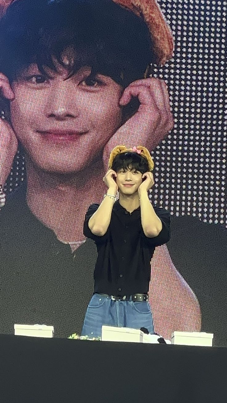 a young man wearing a hat and holding his hands up to his ears while standing in front of a large screen