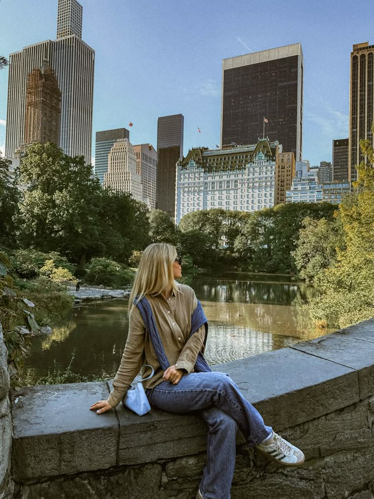 a woman is sitting on a stone wall looking at the water and skyscrapers in the background