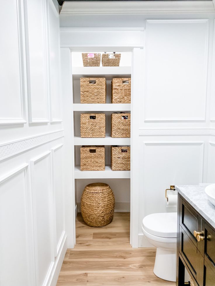 a bathroom with white walls and wooden flooring has baskets on the shelves above the toilet