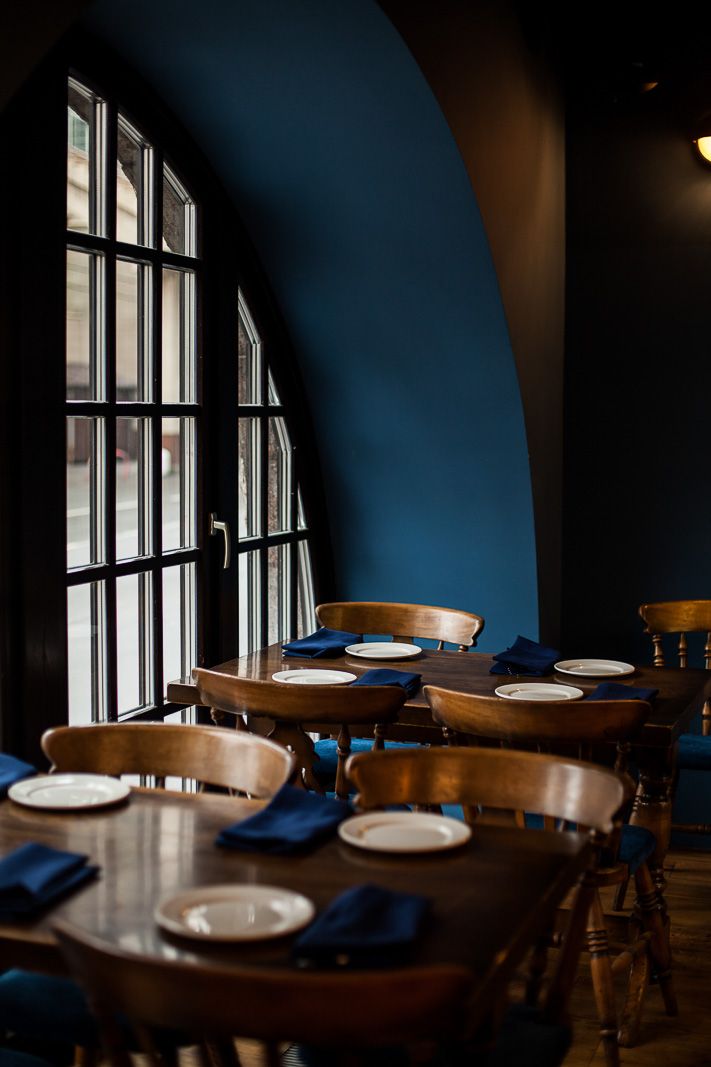 a restaurant with blue walls and wooden tables in front of large arched windows that overlook the street