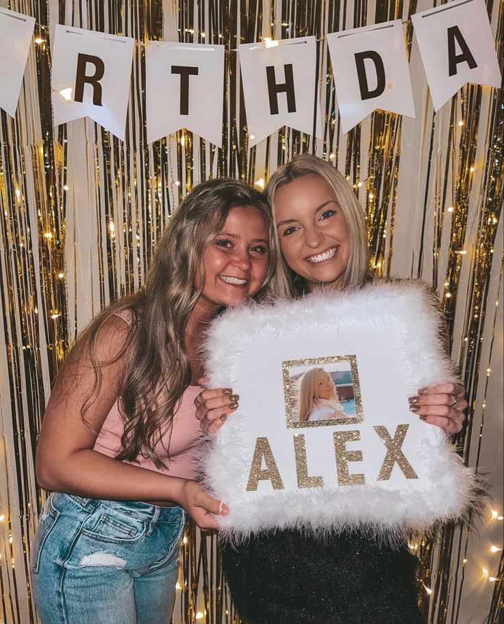 two young women holding up a pillow with the word alex on it in front of a backdrop