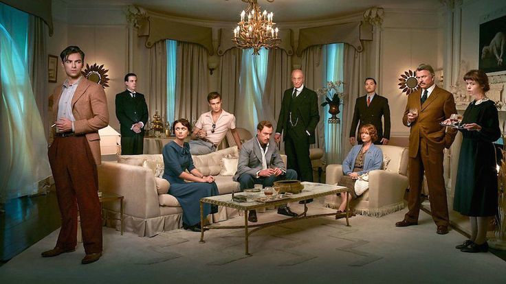 a group of people standing and sitting in a living room next to a chandelier