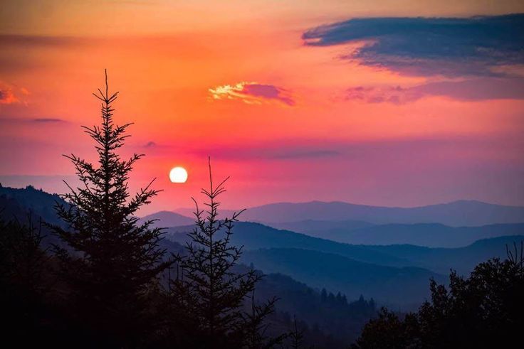 the sun is setting over mountains with trees in foreground and clouds in the background