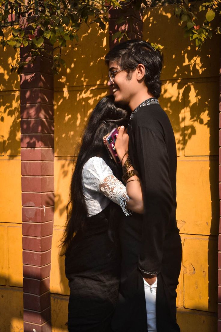 two people standing next to each other in front of a yellow brick wall and tree