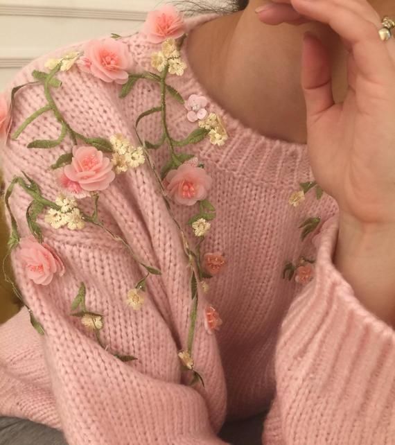 a woman wearing a pink sweater with flowers on the side and holding her hand up to her face