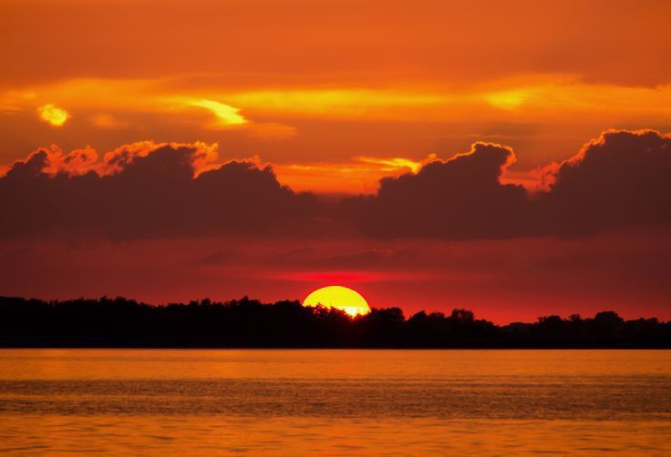 the sun is setting over water with clouds in the sky and trees to the side