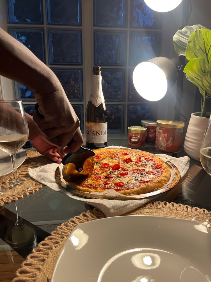 a person cutting a pizza on top of a table next to wine bottles and glasses