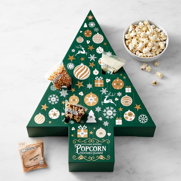 a green christmas tree shaped box filled with popcorn and other holiday treats next to a bowl of candies