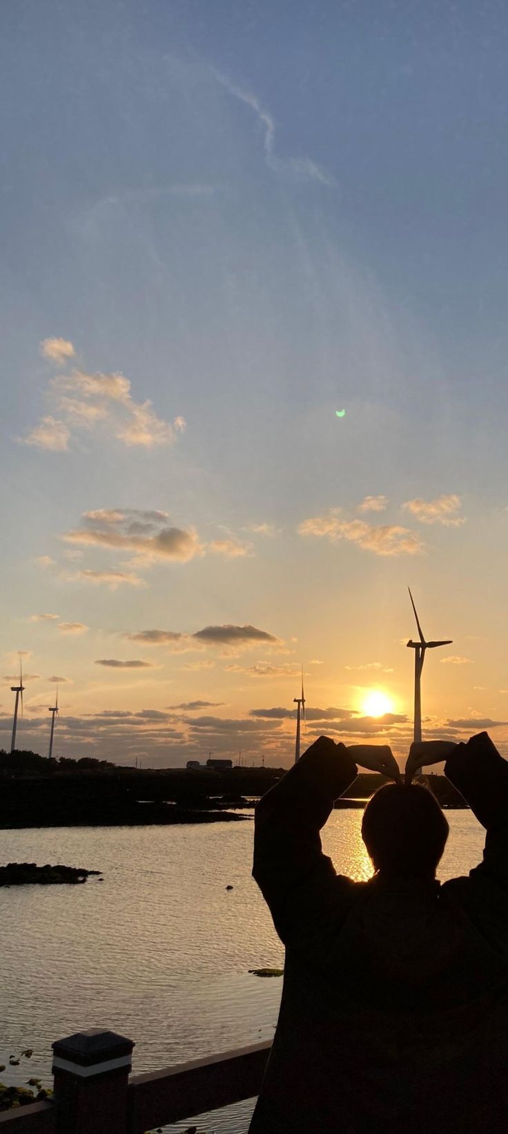 a person taking a photo of the sun setting over water with windmills in the background