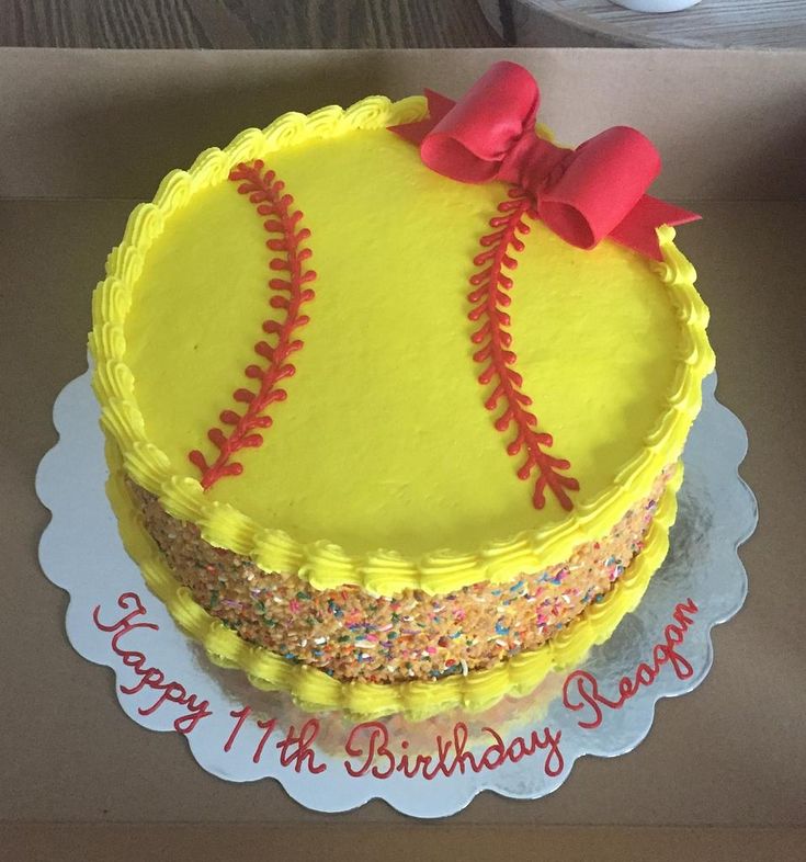 a birthday cake decorated with a softball ball and red bow on top is sitting in a box