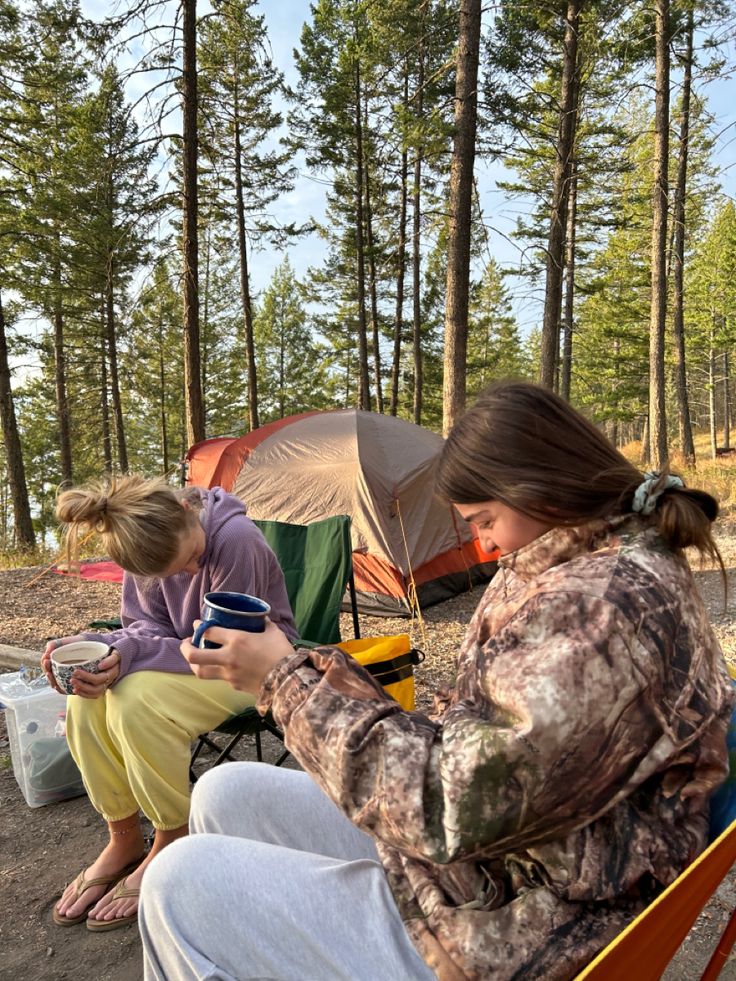 two women sitting on a bench in front of tents and camping equipment, one holding a cell phone