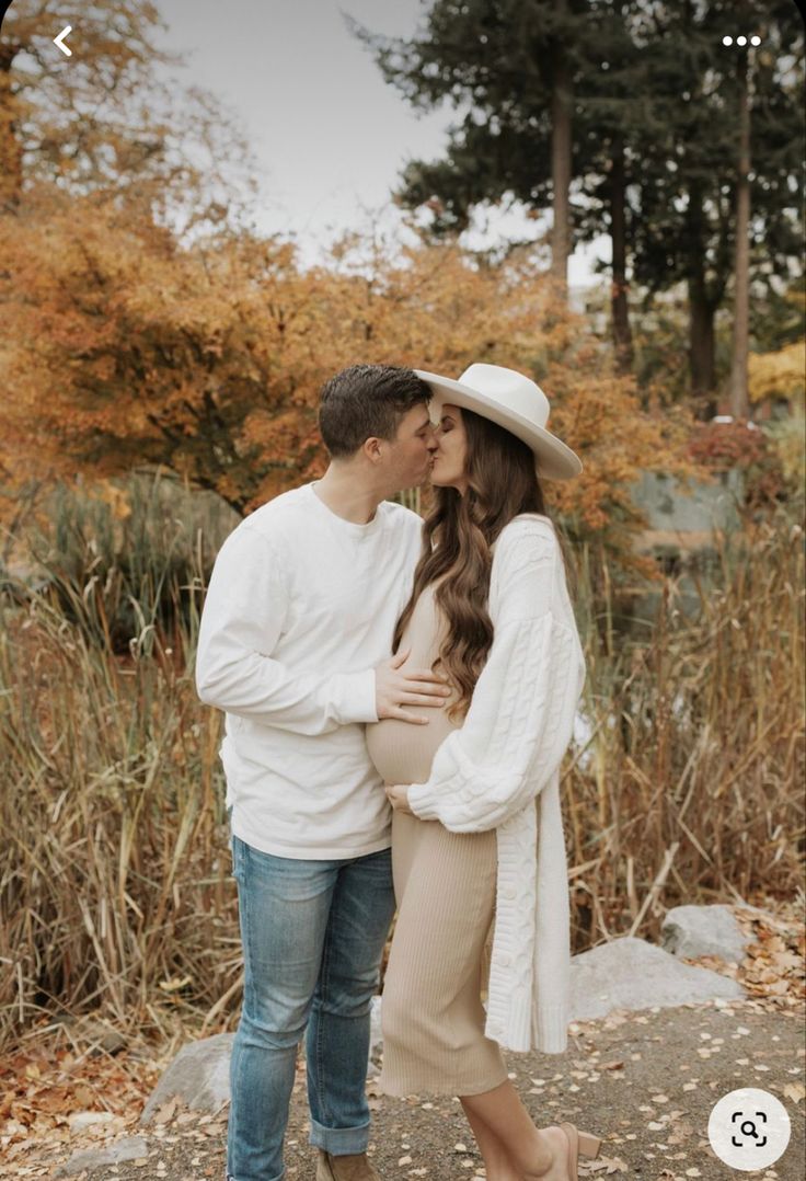 a man and woman standing next to each other in front of trees with fall foliage