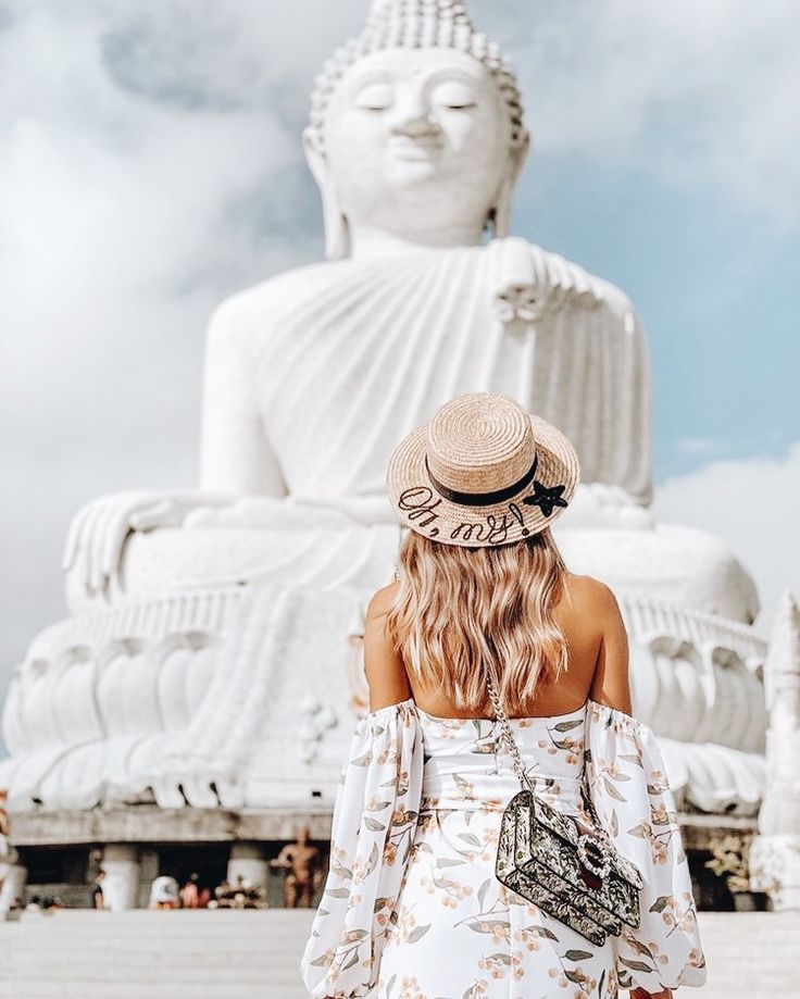 a woman wearing a hat standing in front of a large buddha statue with her back to the camera