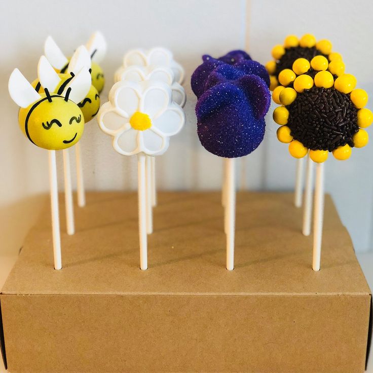 four cake pops decorated with flowers and bees on top of a brown cardboard box against a white wall