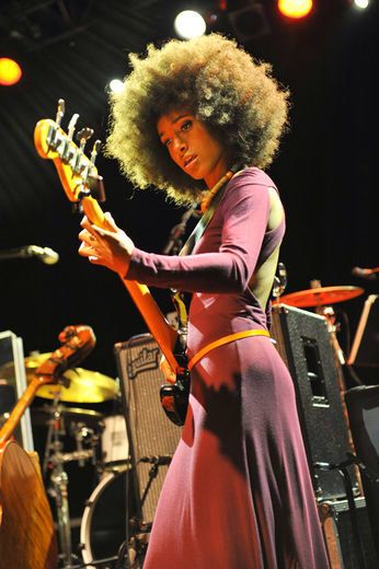 a woman in a purple dress plays an electric guitar on stage with other instruments behind her