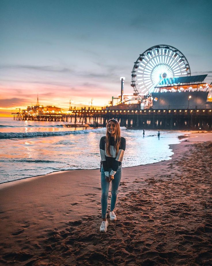 a woman standing on top of a sandy beach next to the ocean at night with a ferris wheel in the background