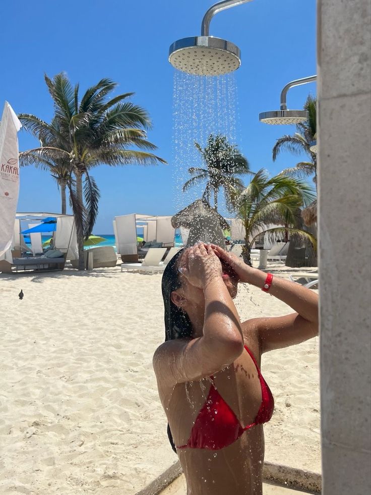 a woman in a red bathing suit standing under a shower head on the beach with palm trees behind her