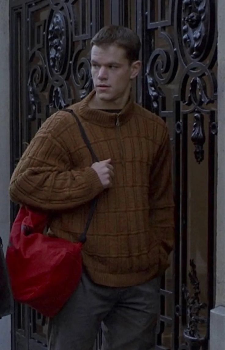 a man with a red handbag is standing in front of a gate and looking at the camera