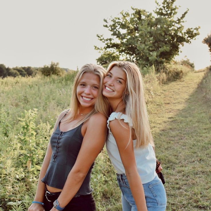 two young women standing next to each other on a field with trees in the background