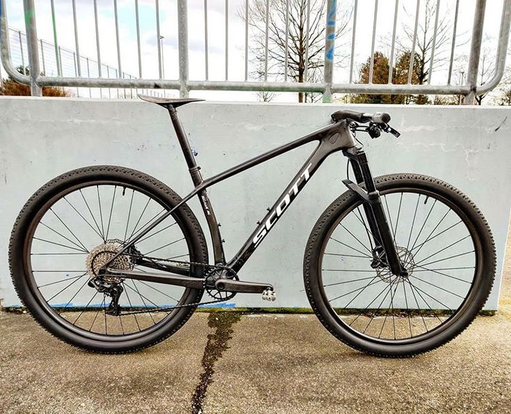 a black bike parked in front of a white wall on concrete ground next to a metal fence