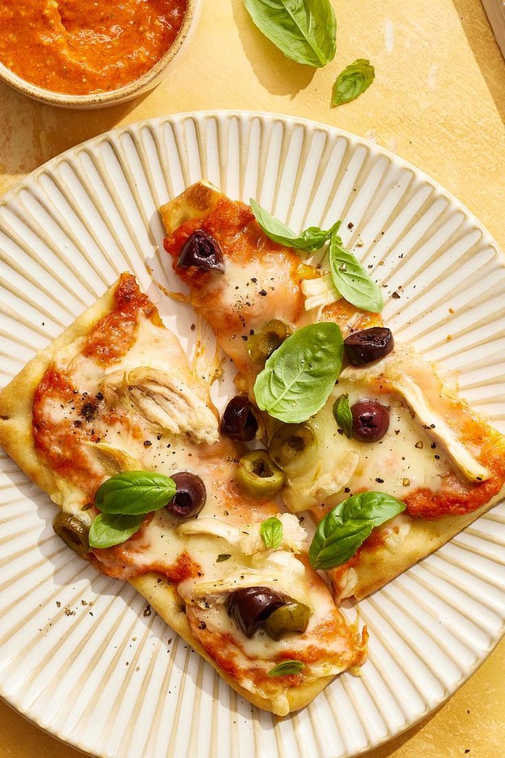 two slices of pizza with olives and basil on a plate next to a bowl of tomato sauce