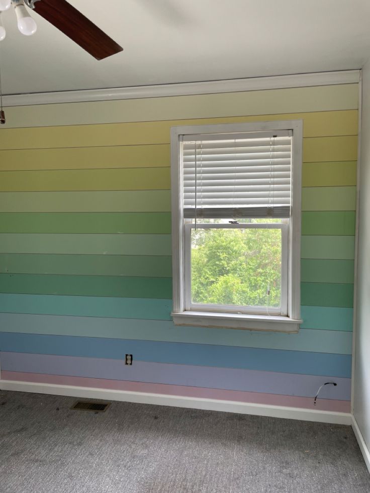 an empty room painted in rainbow colors with a ceiling fan on the far wall and a window