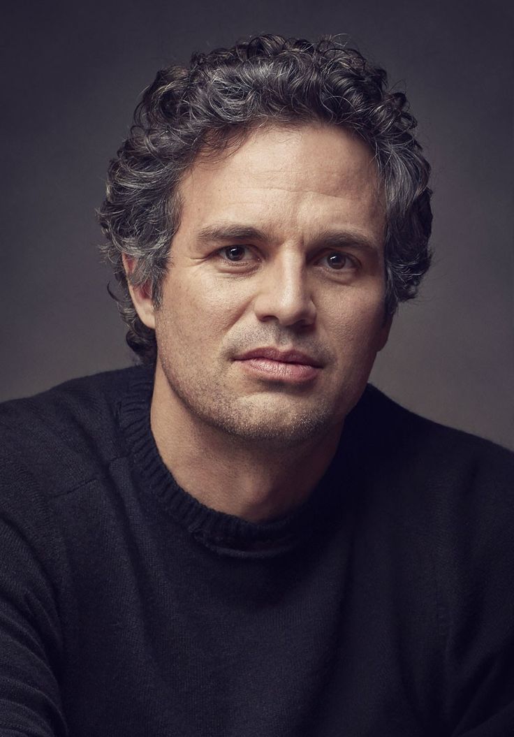 a man with curly hair sitting in front of a dark background wearing a black sweater