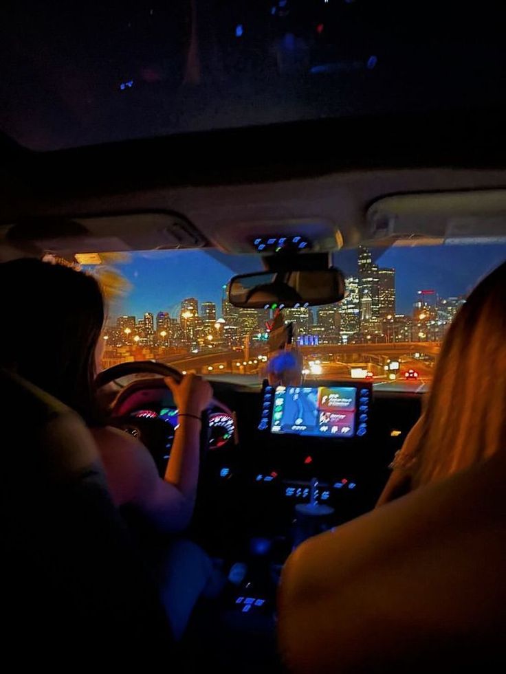 two people in a car driving at night with the city lights lit up behind them