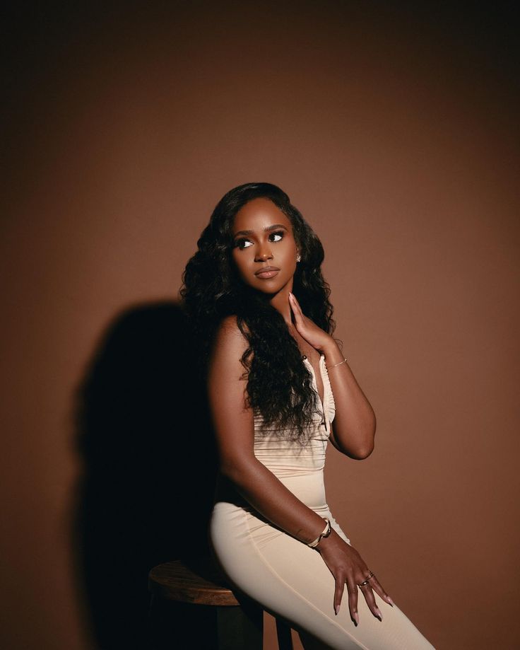 a woman sitting on top of a wooden chair next to a brown wall with her hand under her chin