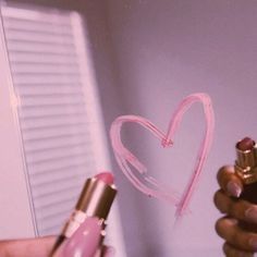 a person holding a pink lipstick in their hand with a heart drawn on the wall behind them