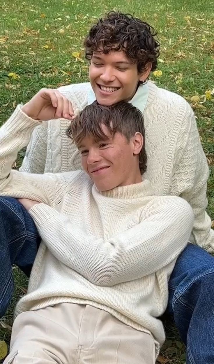 two young men sitting on the ground in front of grass and trees, one with his arm around the other's head