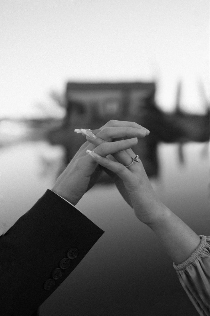 black and white photograph of two people holding hands over each other's fingers with boats in the background