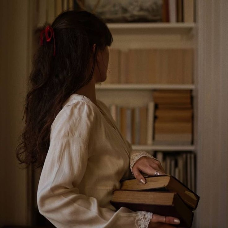 a woman in a white dress is holding two books and looking at the bookcase