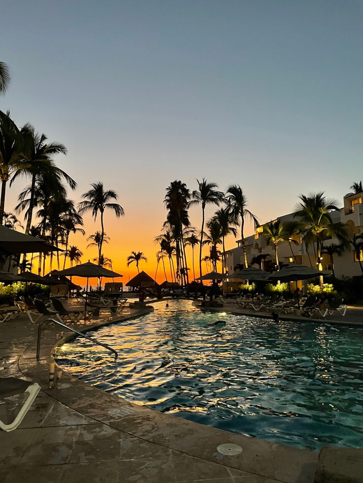 an outdoor swimming pool surrounded by palm trees and chairs at sunset with the sun setting in the background