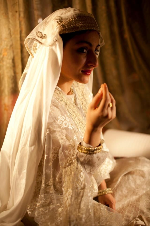 a woman in a white dress and veil sitting on a bed with her hands together