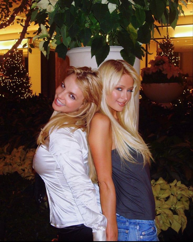 two beautiful young women standing next to each other in front of a potted plant