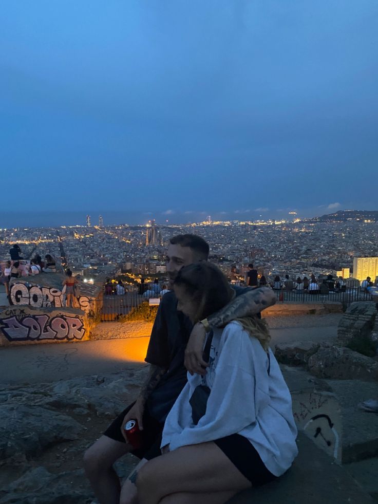 a man and woman sitting on top of a rock with the city lights in the background